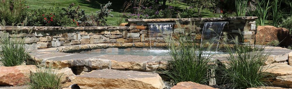 Looking for Landscaping Design Near me - Contact Burkholder Landscaping - Retaining wall surrounding pool with waterfall and surrounded by softscape plants and trees and rocks