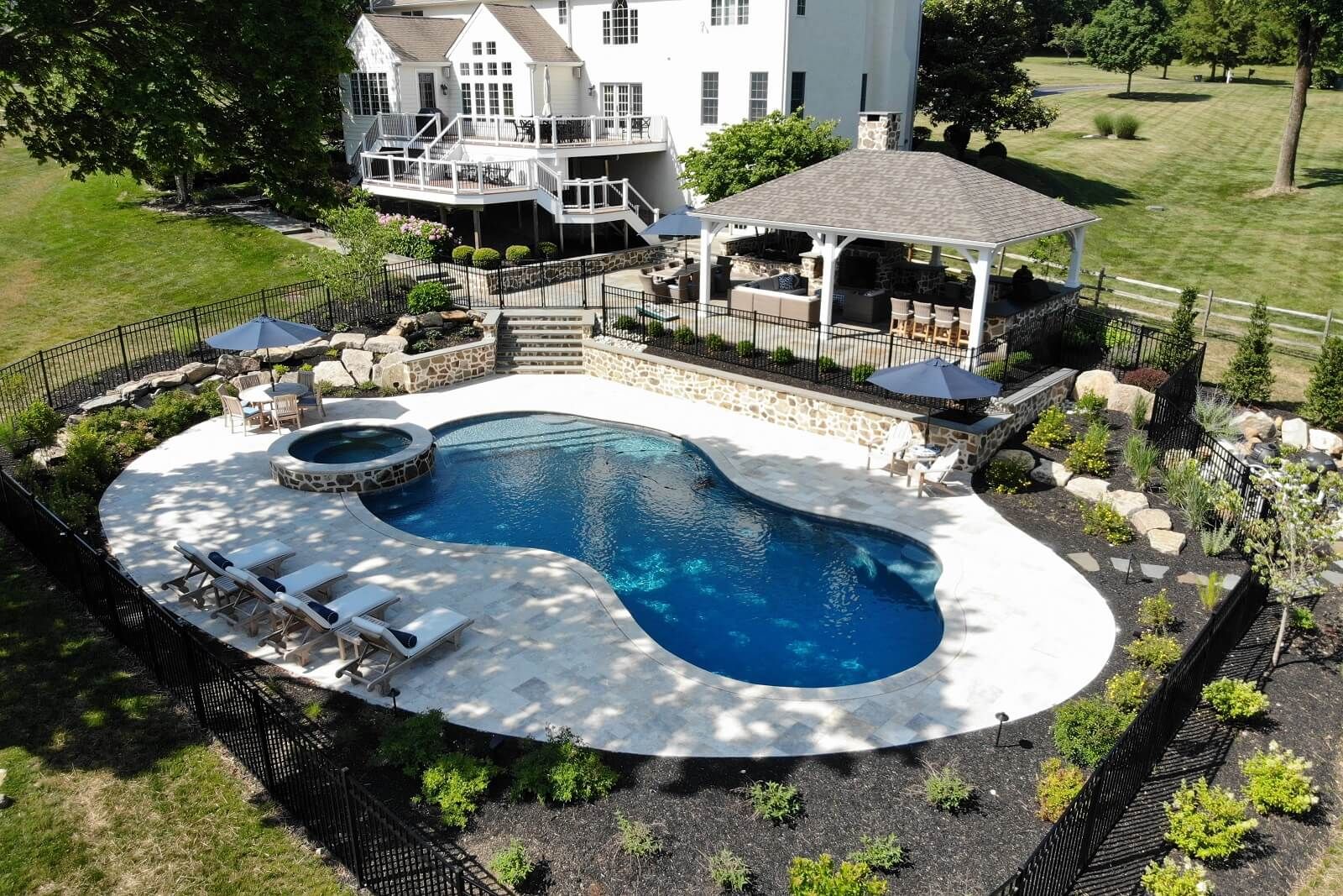 Backyard of a large white house with double layer deck, walkway to pavilion and pool with spa - Burkholder Landscape