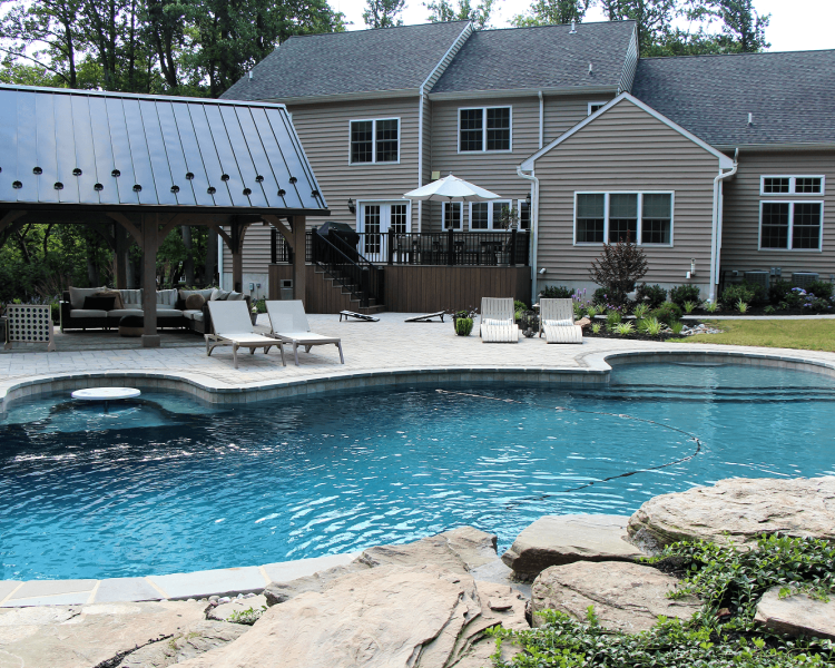 Backyard of a tan house with a deck. A pool surrounded by a light colored deck with chaises, pavilion with furniture -Burkholder Landscape