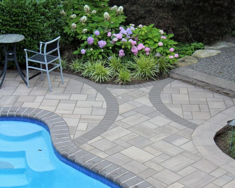 Edge of a blue pool surrounded by a tiled walkway and lush foliage-Burkholder Landscape