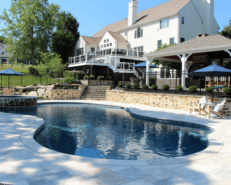Pool surrounded by a light colored walkway with a spa, retaining wall with steps up to pavilion and a multilevel white deck -Burkholder Landscape