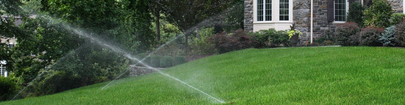 sprinklers running on green lawn by a home and wooded area | How to Care for Your Lawn during Drought Conditions | Burkholder Landscape
