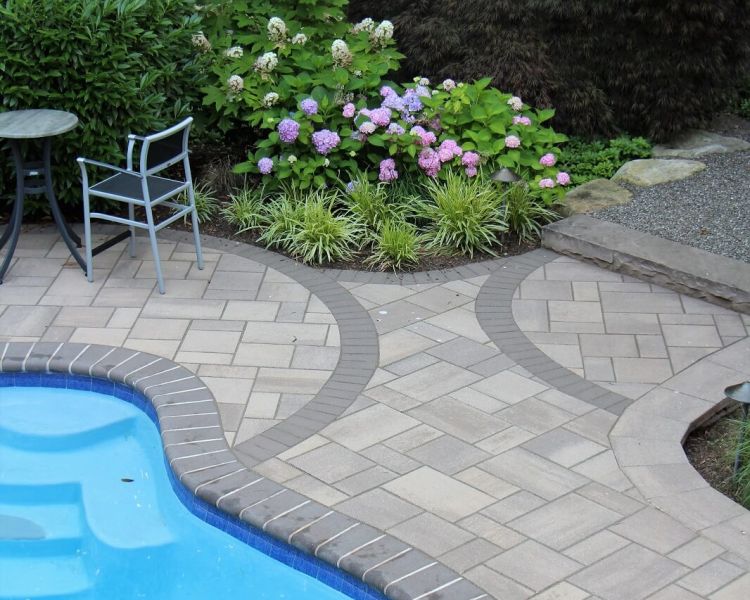 Edge of a blue pool surrounded by a tiled walkway and lush foliage-Burkholder Landscape